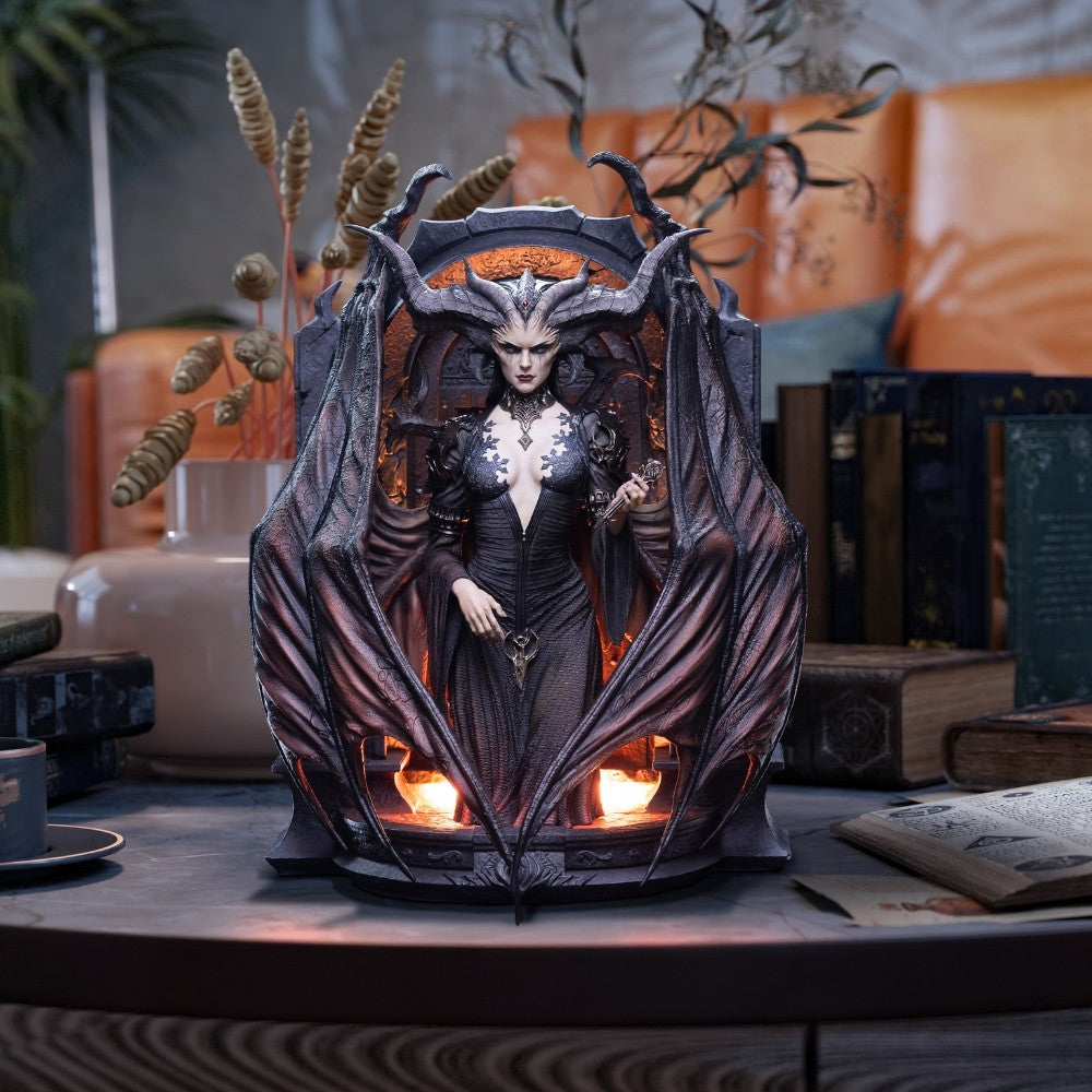 Diablo IV – Lilith Art Statue (HEX Collectibles). A gothic fantasy figurine of a horned female demon in a black dress, seated within a bat-winged throne structure with glowing candles, displayed on a tabletop in a styled room setting.