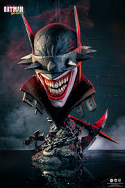 Batman Who Laughs - 1/1 Scale Art Mask (PureArts). Batman Wicked: The Joker statue bust with a wide, menacing grin, dark textured mask, and red accents, displayed on a reflective base against a smoky dark brick background. Batman Wicked logo is visible in the top left.