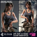 Lara (Tomb Raider) Realistic Silicone 1:1 Replica Cosplay Model Doll (Game Lady). Promotional product image for a life-size 1:1 scale silicone human replica of Lara Croft from Tomb Raider, shown in two side-by-side views. The left view shows the character against a dark gray background with text stating 'Tomb Raider "Lara Croft" Cosplay Outfit Included,' and the right view shows her in a rainier outdoor scene. Lara Croft Sex Doll.